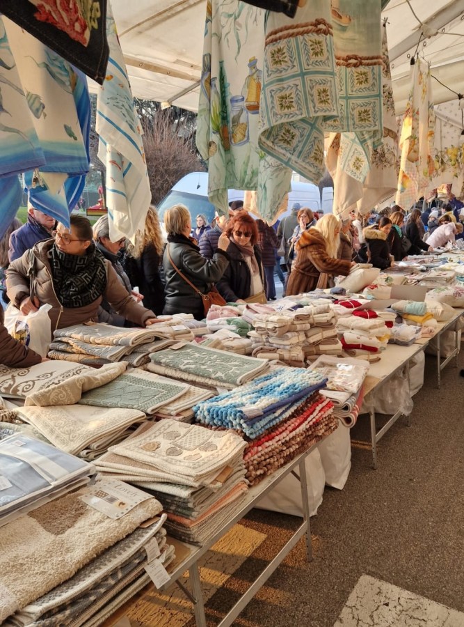 Mercato di qualità con bancarelle di tessuti, asciugamani e biancheria per la casa, frequentato da un pubblico numeroso in un'area coperta.