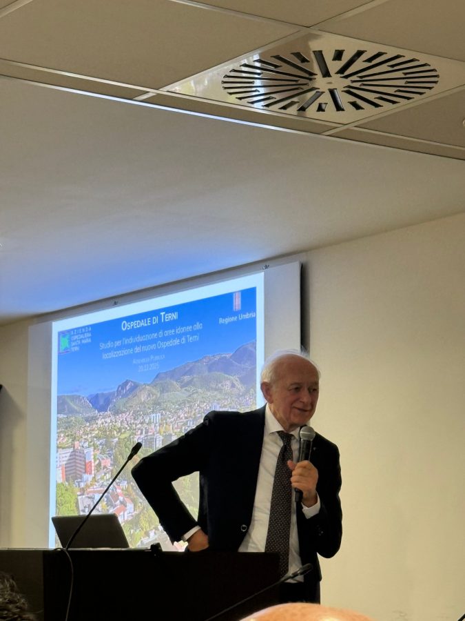 Ingegner Binini parla durante la presentazione dello studio sulla localizzazione del nuovo ospedale di Terni, con uno schermo che mostra il titolo della presentazione sullo sfondo.