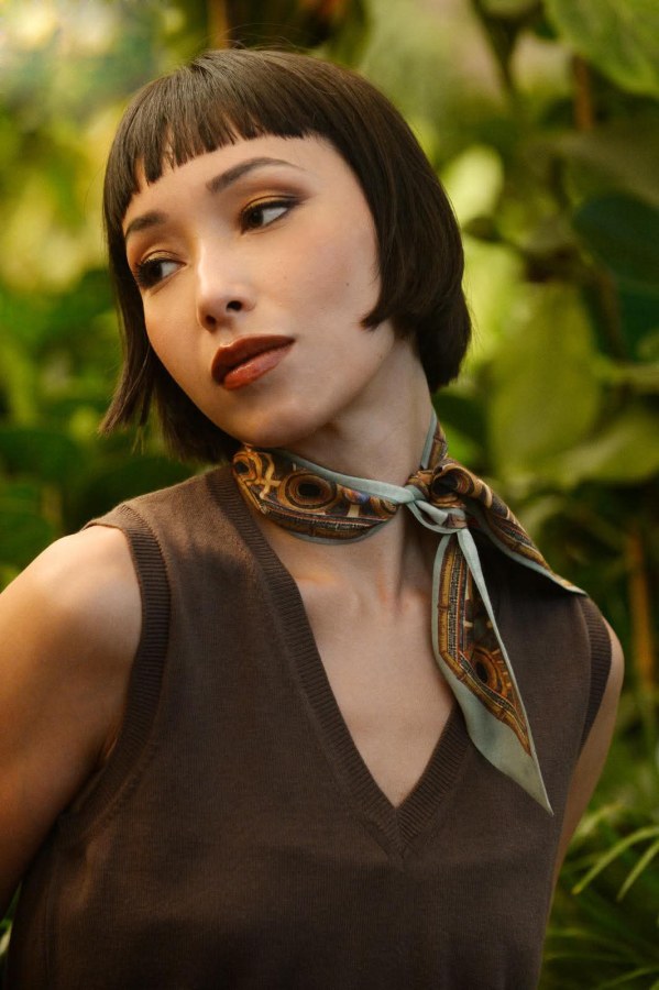 Ritratto di una giovane donna con capelli corti e castani, che indossa un top senza maniche marrone e una sciarpa di seta intorno al collo, con uno sfondo verde e vegetazione.