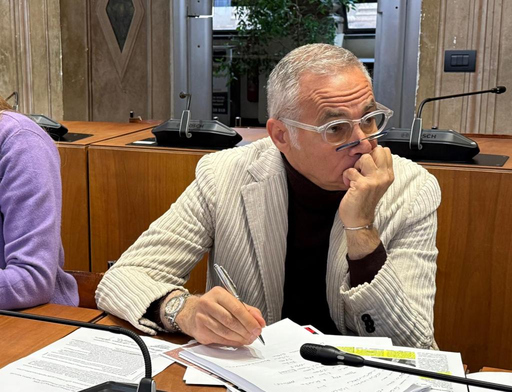 A thoughtful individual with glasses and a striped jacket sits at a table, hand on chin, surrounded by papers and a microphone.