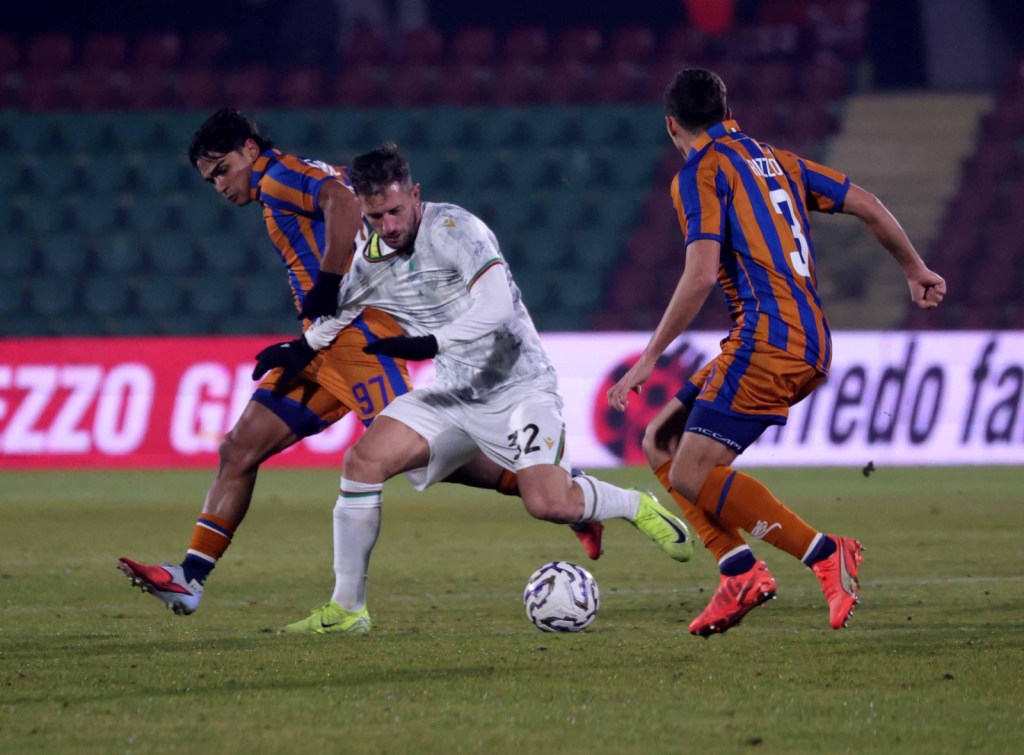 A moment from a soccer match showing two players in orange and blue jerseys challenging a player in white, with an audience seen in the background.