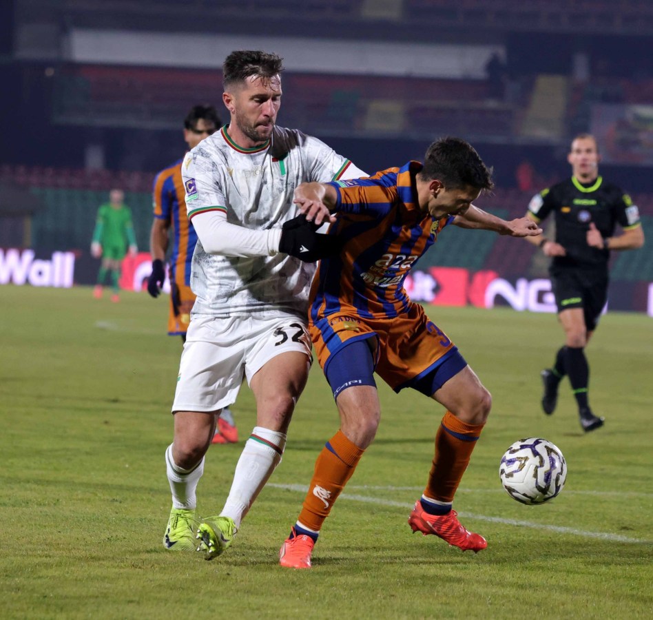 A football match scene featuring two players in action, one wearing a white kit with the number 32 and the other in an orange and blue striped kit, competing for the ball on a green pitch.