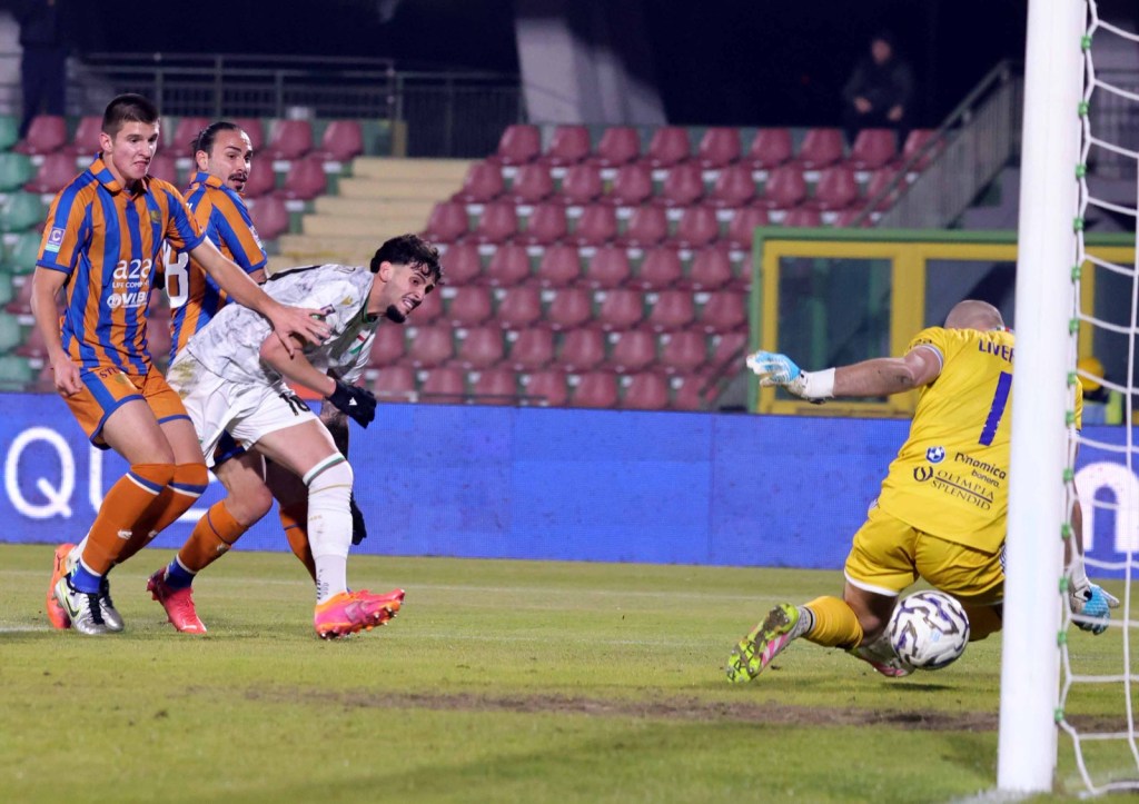 A dramatic moment in a soccer match showing a player celebrating a near goal, while a goalkeeper dives to prevent it, with teammates in the background.