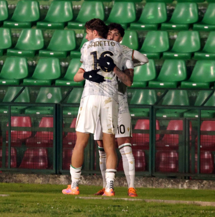 Two football players embrace on the field, wearing jerseys with the numbers 16 and 10. They are standing in front of green stadium seating.