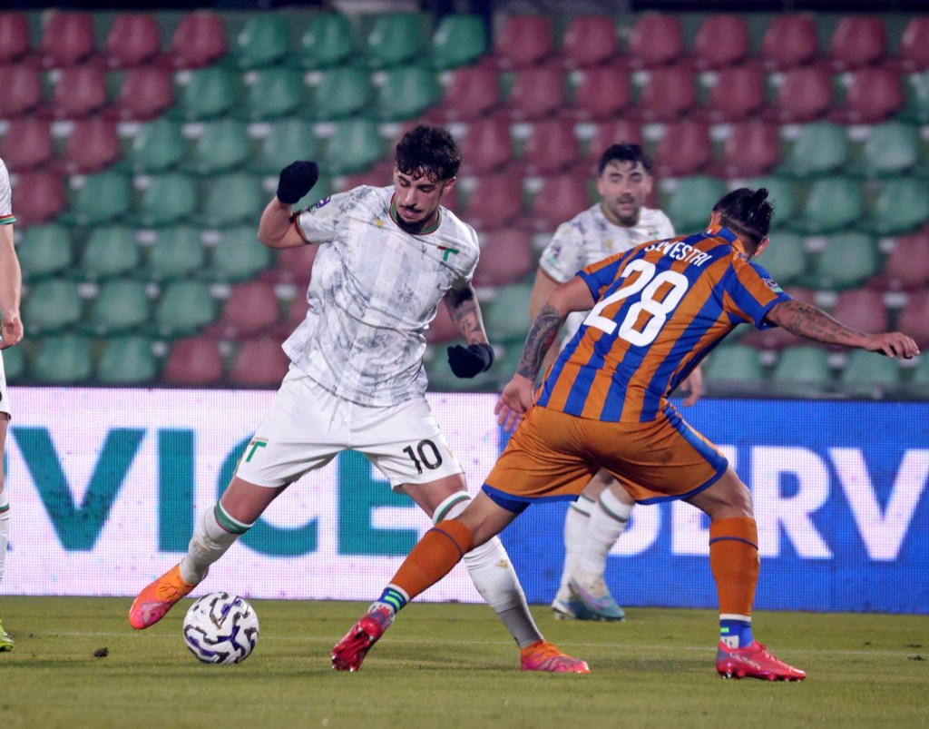 Two soccer players competing for the ball on a field. One player in a white jersey with the number 10 is attempting to evade a challenge from another player wearing an orange and blue striped jersey with the number 28.