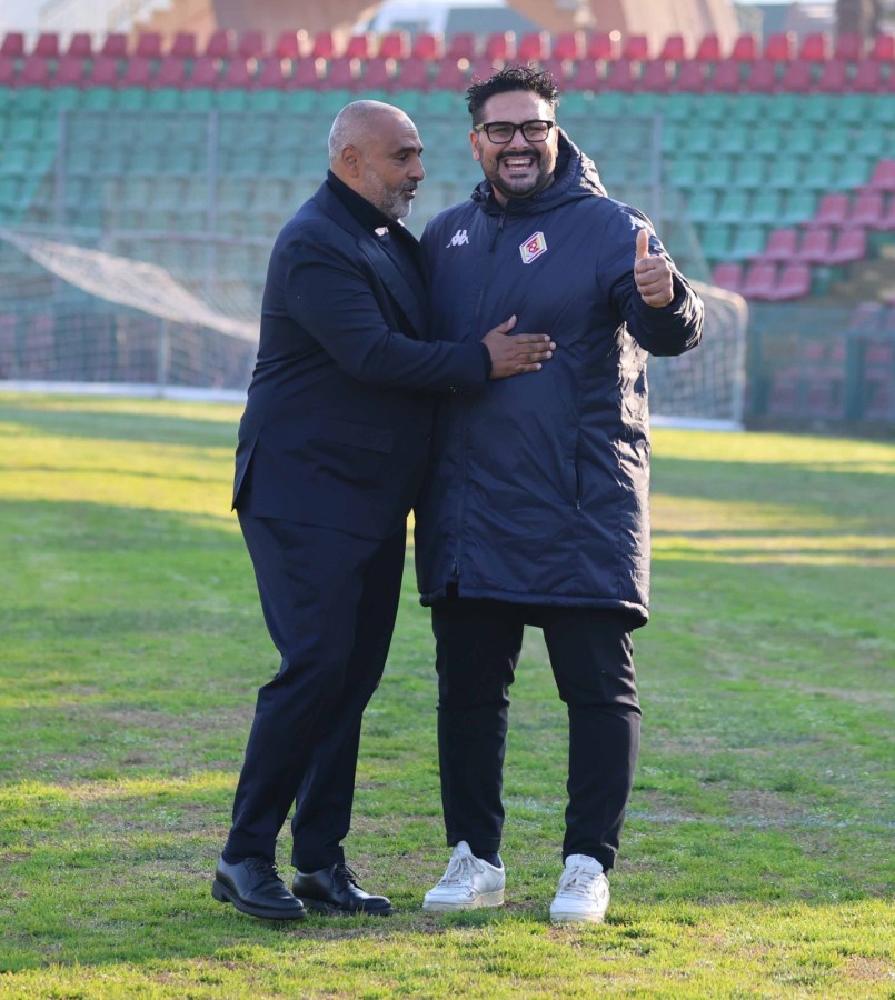 Two men smiling and posing together on a football field, one in a suit and the other in a sports jacket, with a green and red stadium seating in the background.