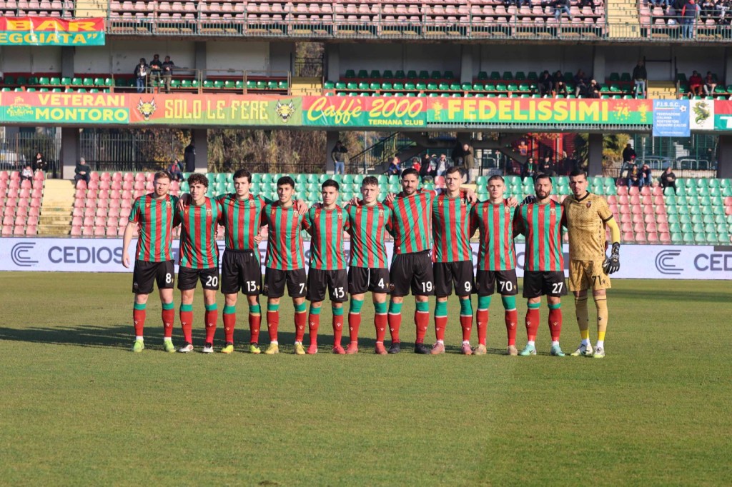 Football team lined up for a match, wearing striped red and green jerseys, with a goalkeeper in beige uniform, standing on the field in a stadium.