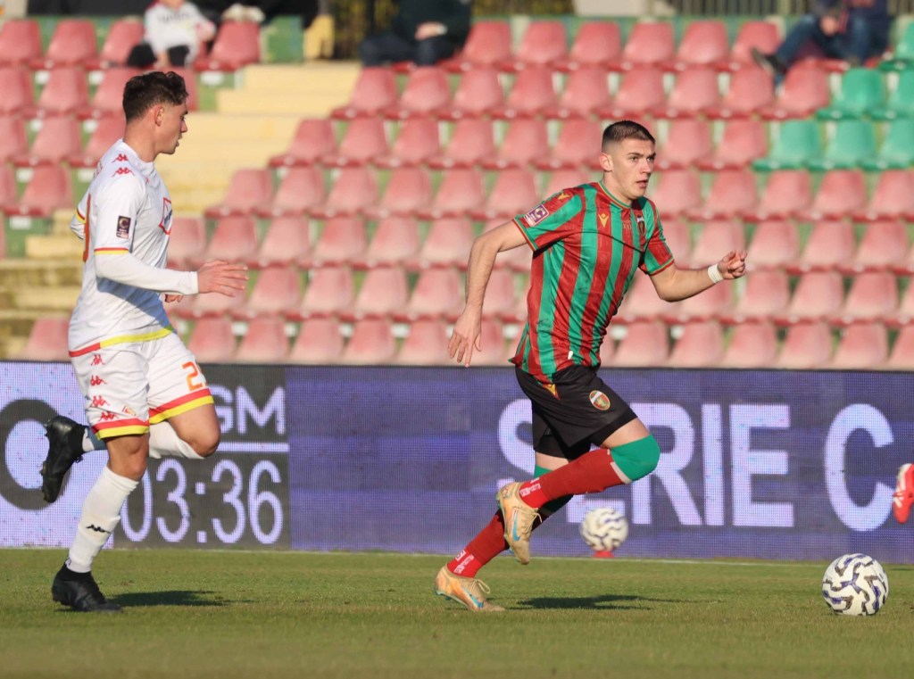 Two soccer players compete on the field, one in a green and red striped jersey and the other in a white jersey with yellow and red accents, set against a backdrop of empty stadium seats.
