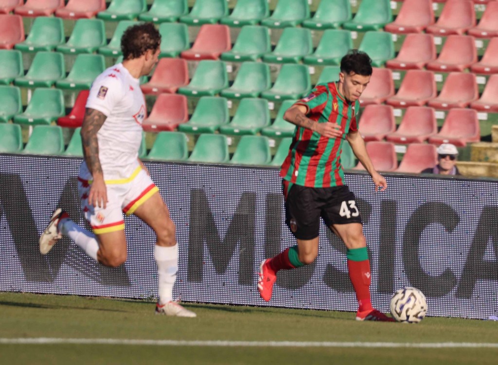 A soccer player wearing a red and green striped jersey dribbles the ball on a field, while another player in a white jersey trails behind.