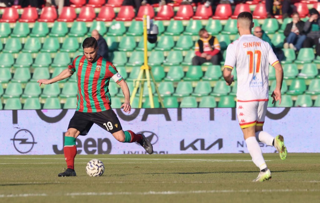 A soccer player in a green and red striped jersey, wearing number 19, is kicking a soccer ball on the field. Another player, wearing a white jersey with the name 'SINANI' and number 11, is running nearby. The background features a stadium with green and red seating.