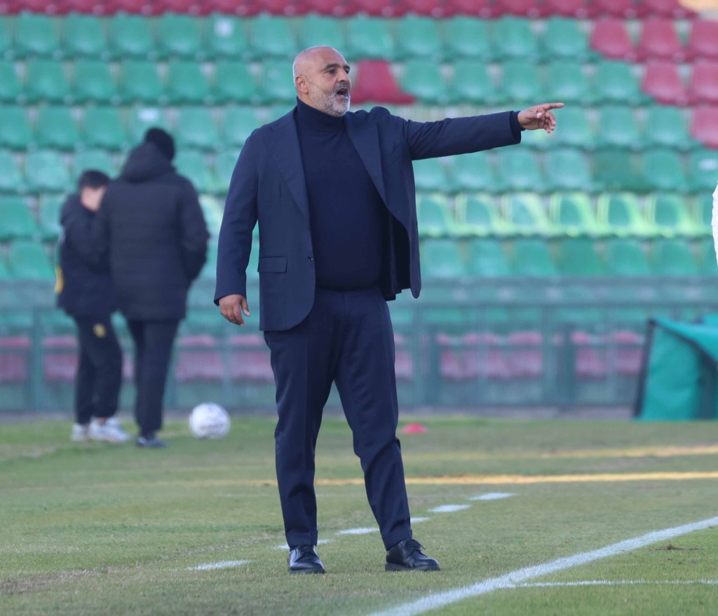 A football coach dressed in a suit confidently points while giving instructions on the field, with empty stands in the background.