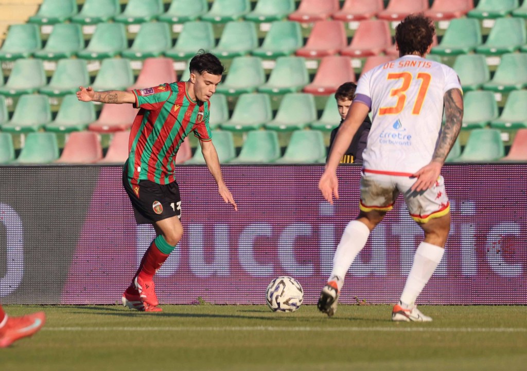 A soccer player in a green and red striped jersey dribbles the ball on the field while another player in a white jersey with the number 31 looks on.
