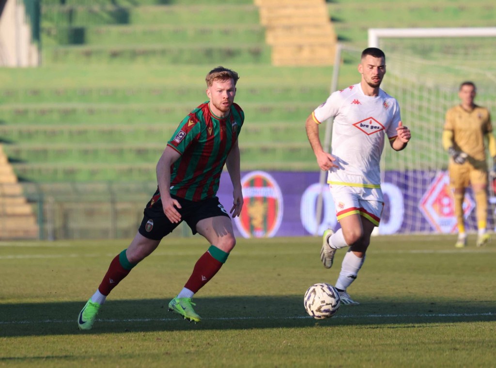 A soccer match in progress featuring two players: one wearing a green and red striped jersey and the other in a white jersey. The background shows empty bleachers and a soccer goal.