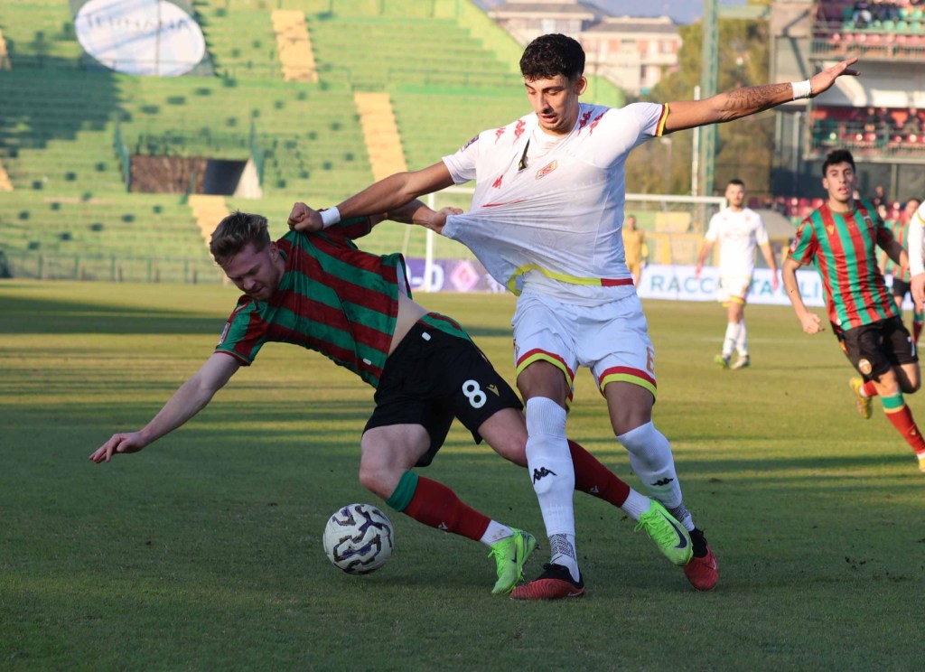 A soccer match scene where a player in a red and green striped jersey is falling while being challenged by an opponent in a white jersey, with players in the background.