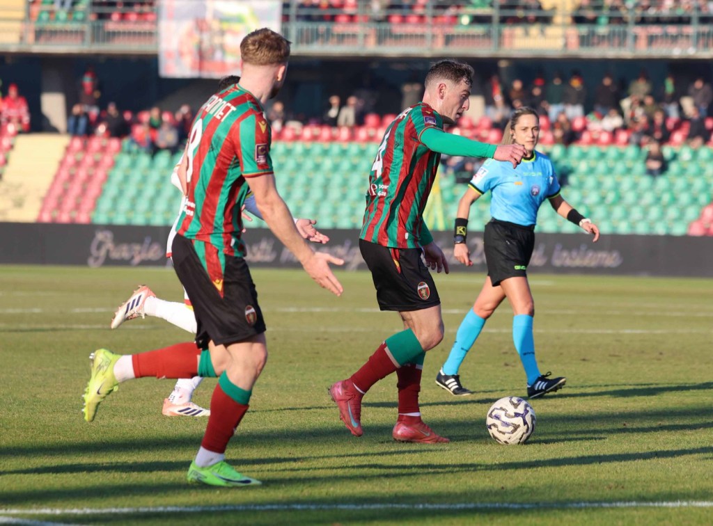 Two soccer players wearing green and red striped jerseys on a field, with a referee in blue observing the play. A soccer ball is in the foreground.