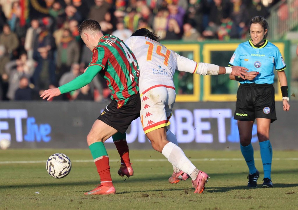 A soccer match action shot featuring two players competing for the ball, one in a green and red striped jersey and the other in a white jersey with red accents. An official in a blue uniform observes the play.