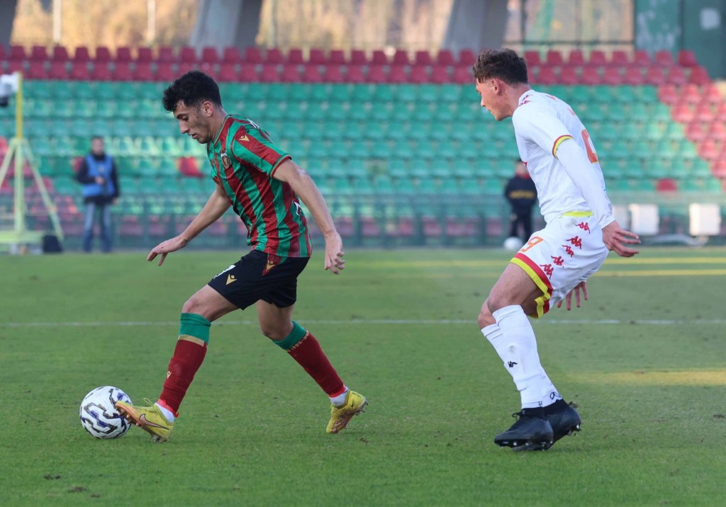 A soccer match featuring two players; one in a green and red striped jersey dribbling the ball while the other in white attempts to intercept.