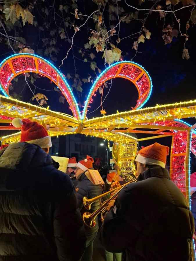 Un gruppo di musicisti in abiti invernali suona strumenti musicali sotto un'arco di luci natalizie, indossando cappelli di Babbo Natale durante la parata natalizia.
