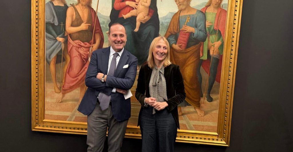 Due persone sorridono davanti a un dipinto, indossando abiti eleganti, all'interno di una galleria d'arte.