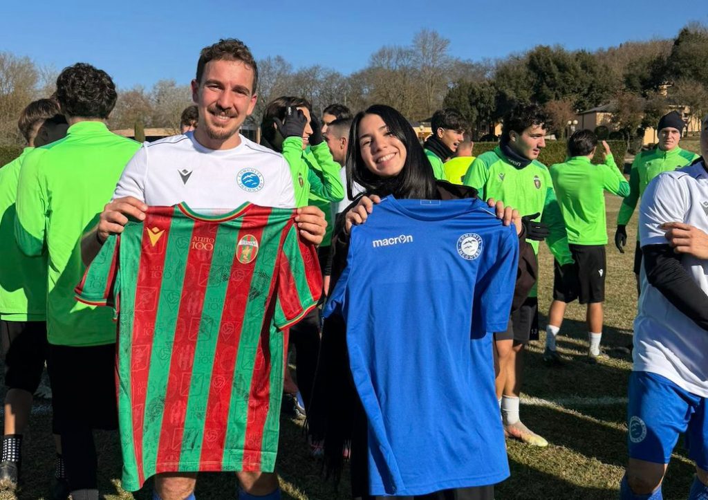 Due persone sorridenti, un uomo e una donna, tengono in mano le maglie di calcio delle rispettive squadre. Sullo sfondo, un gruppo di giovani calciatori in maglie verdi e bianche si prepara per una sessione di allenamento.