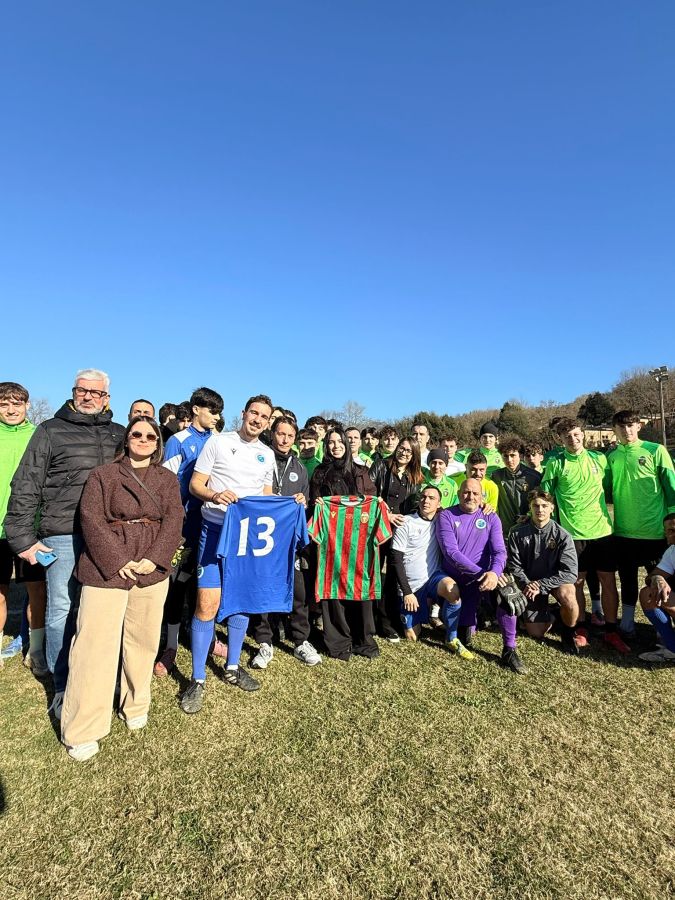 Gruppo di giovani calciatori e membri della Comunità Incontro posano insieme su un campo di calcio, mostrando maglie da gioco. Sullo sfondo, un cielo blu e colline.