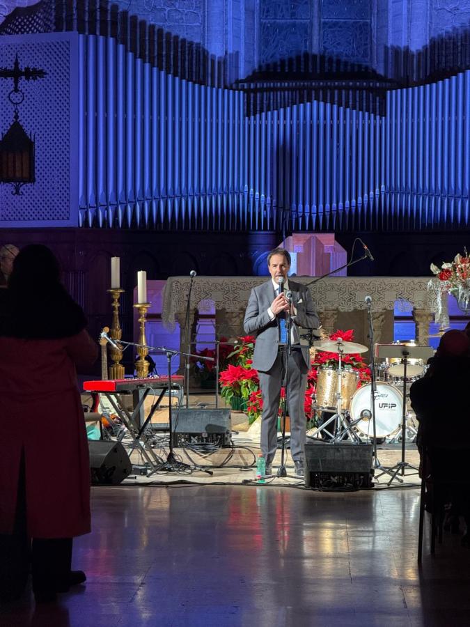 Un uomo in giacca e cravatta parla al microfono durante un concerto gospel, con un palco allestito e decorazioni floreali sullo sfondo.