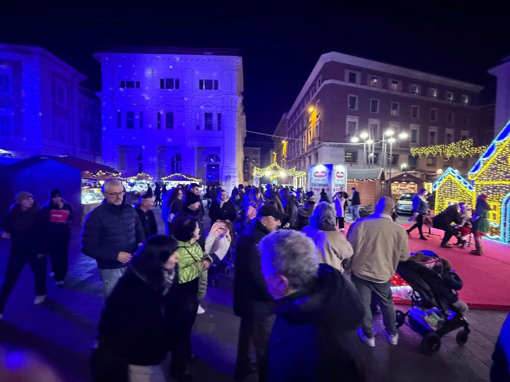 Un gruppo di persone in una piazza illuminata da luci festive, con decorazioni natalizie e bancarelle, durante la notte.