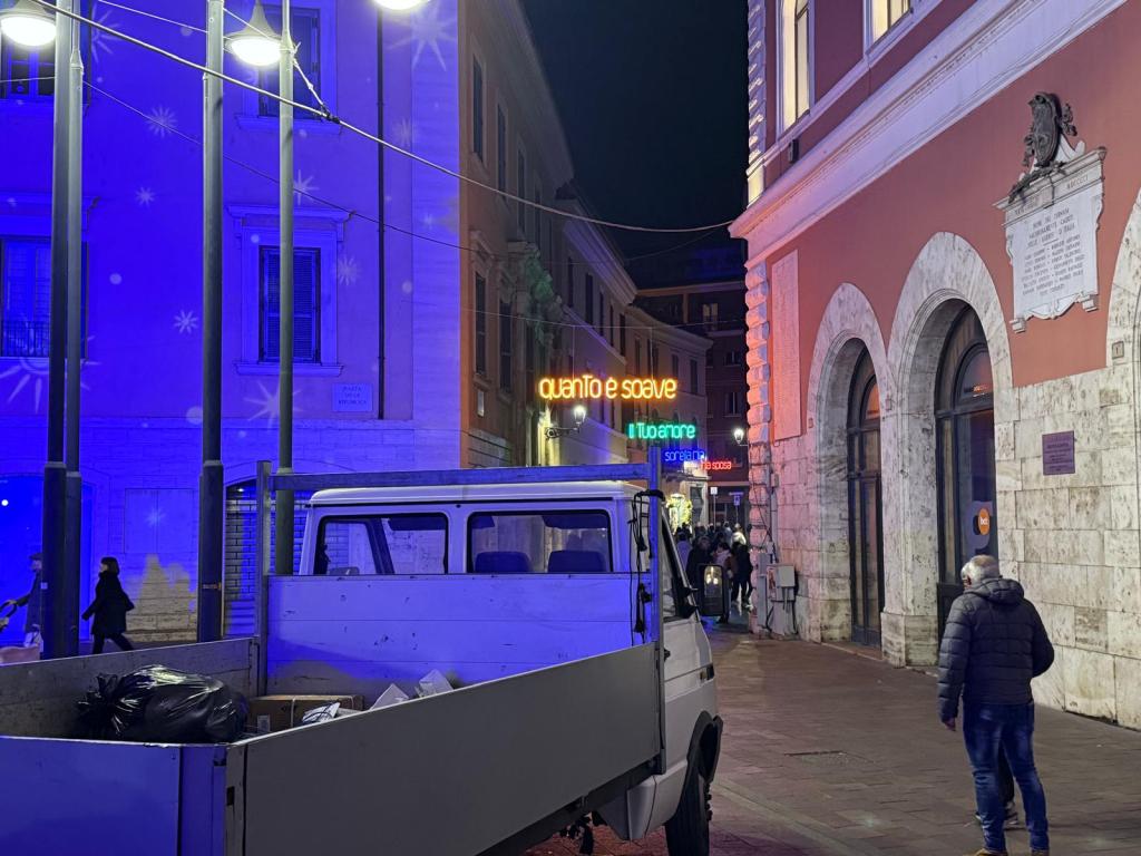 Una strada illuminata di notte con un furgone bianco e decorazioni luminose visibili sugli edifici circostanti.