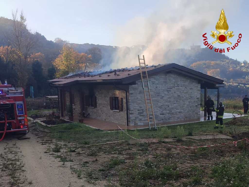 Una casa di pietra coinvolta in un incendio controllato, con i vigili del fuoco al lavoro e fumi che si sollevano dal tetto.