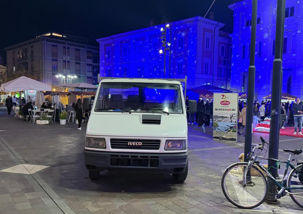 Un furgone Iveco parcheggiato in una piazza affollata di notte, con luci blu che illuminano gli edifici circostanti e stand di cibo visibili sullo sfondo.