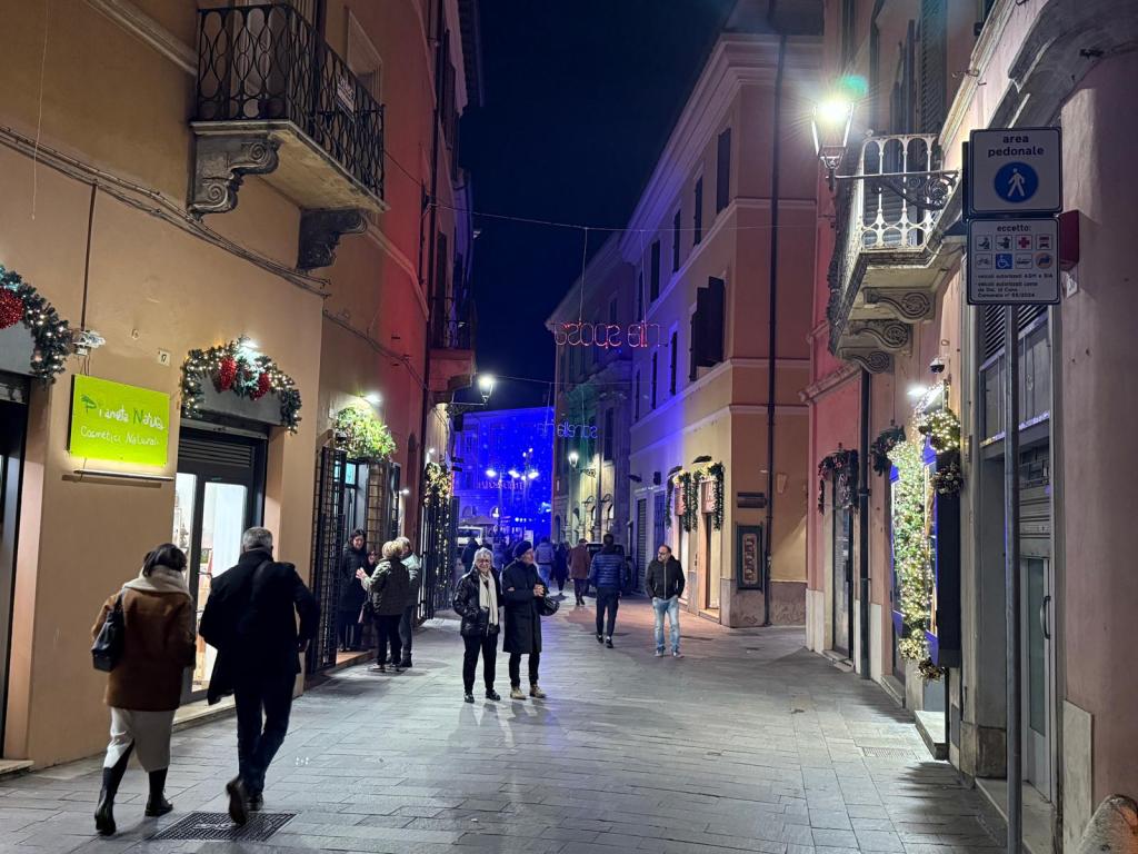Una strada illuminata di notte, decorata con addobbi natalizi. Persone passeggiano lungo il marciapiede, con negozi e luci festive sullo sfondo.