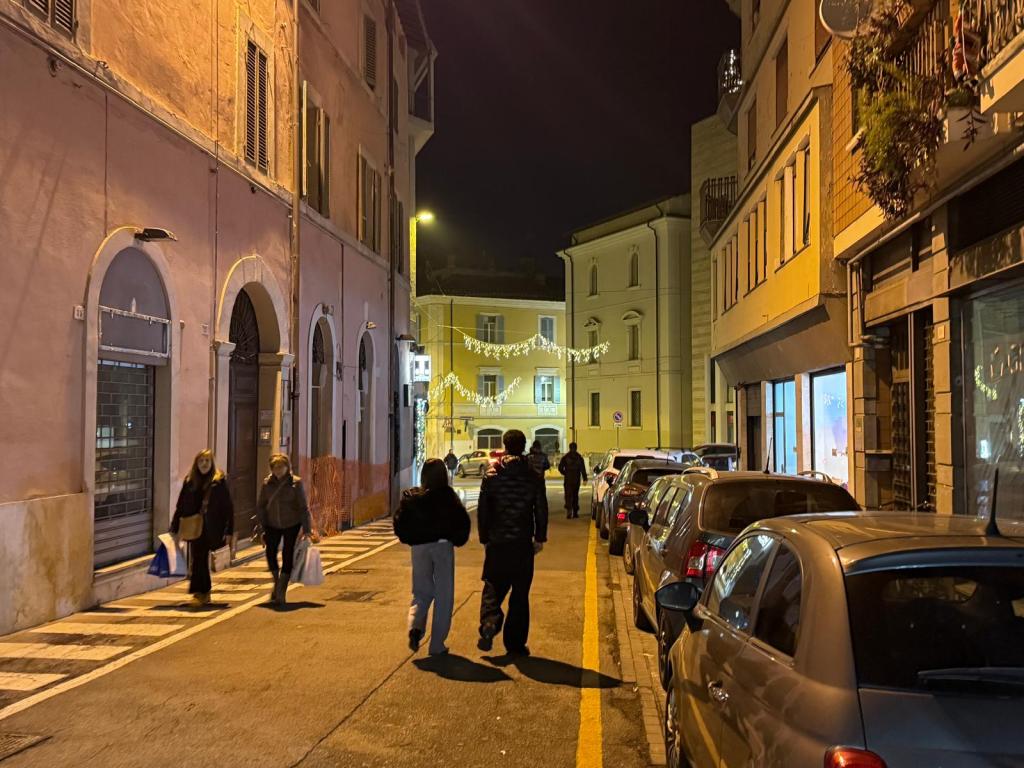 Una strada urbana illuminata di notte con persone che camminano e auto parcheggiate lungo il marciapiede. Edifici colorati e decorazioni luminose visibili in background.
