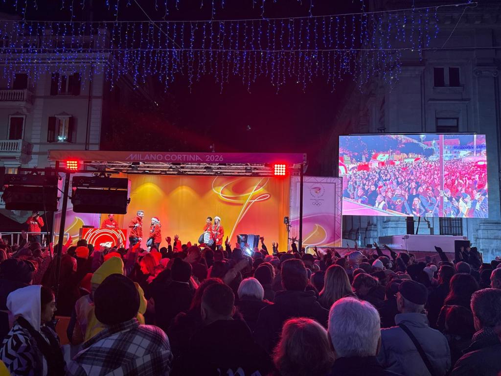 A lively crowd gathers at an outdoor event celebrating the Milano Cortina 2026 Olympics, with performers on stage and colorful lights illuminating the scene.