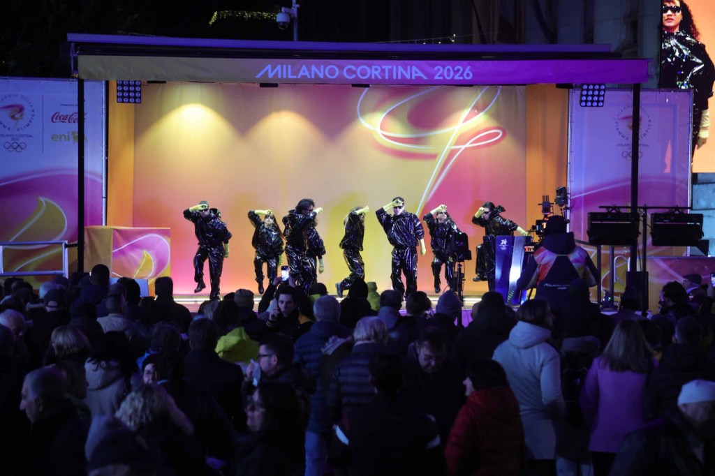 A crowd watches a dance performance on a brightly lit stage decorated for the Milano Cortina 2026 event, with performers in black outfits and colorful lighting.