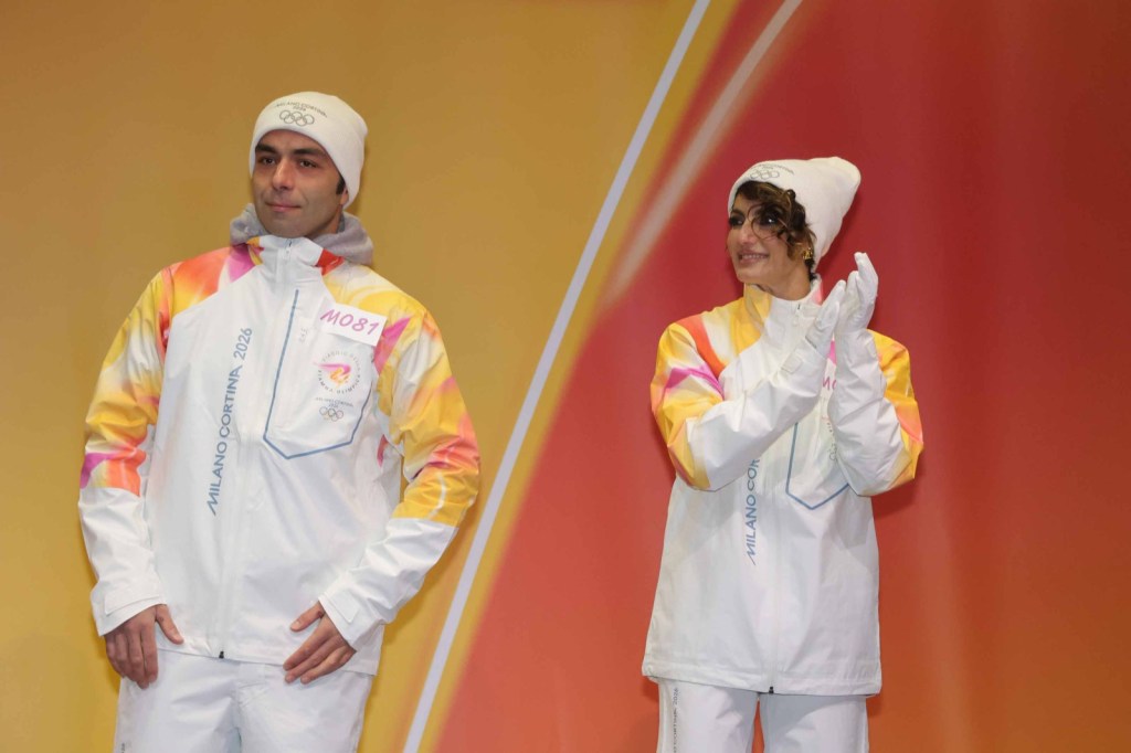 A male and a female athlete are wearing colorful winter jackets with Olympic branding, posing in front of an orange background at an event promoting the Milano Cortina 2026 Olympics.
