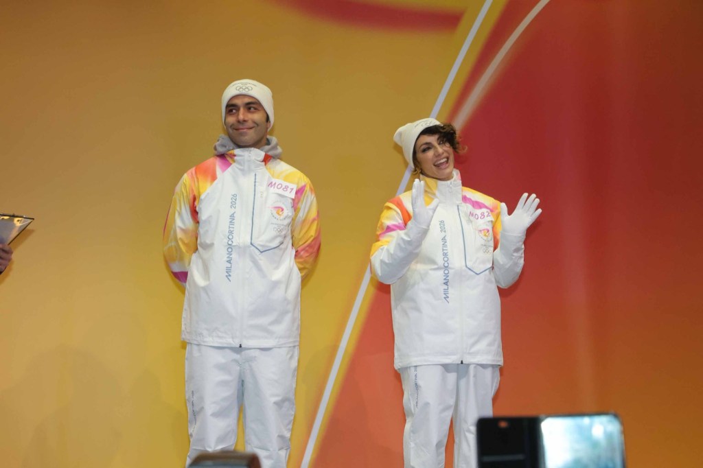 Two athletes on stage, smiling and wearing white sports jackets with colorful designs, promoting the Milano-Cortina 2026 Olympics.