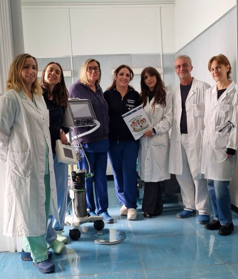 A group of seven medical professionals standing together in a hospital setting, wearing white lab coats and medical scrubs. They are smiling and holding a publication, with medical equipment visible in the background.