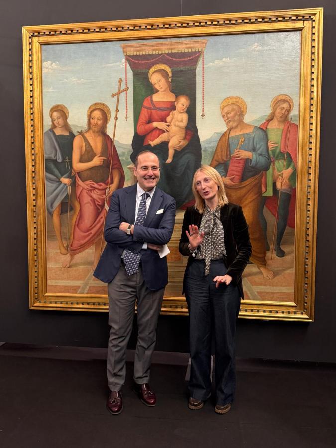 Two individuals standing in front of a large, ornate painting depicting Madonna with child and several saints.