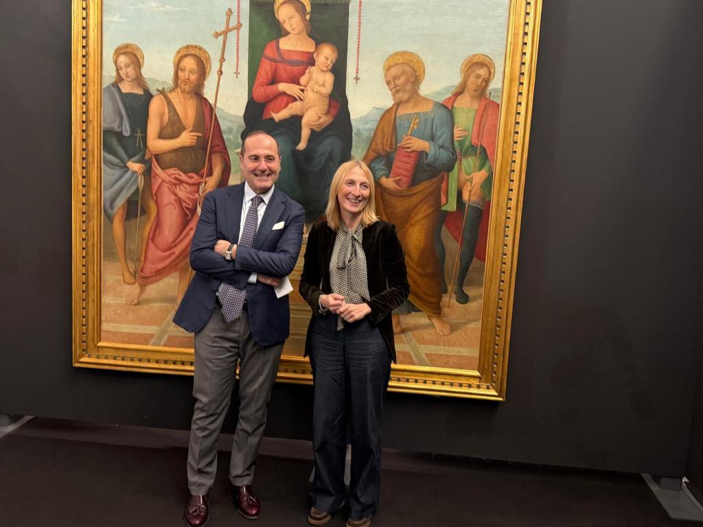 Two individuals posing in front of a religious painting featuring figures from the Christian tradition, including Mary and Jesus, with a gold frame.