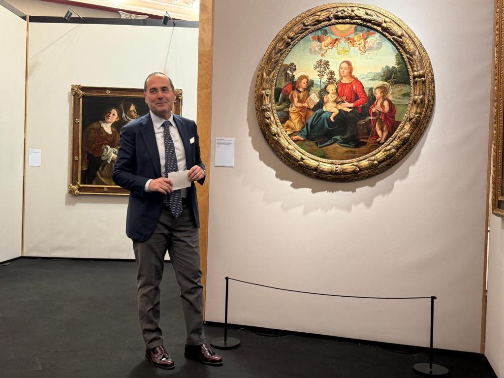 Un uomo in abito formale sta in piedi accanto a un dipinto rotondo che rappresenta la Madonna con Bambino, circondato da figure angeliche. L'ambiente è un'esposizione d'arte con opere incorniciate visibili sullo sfondo.