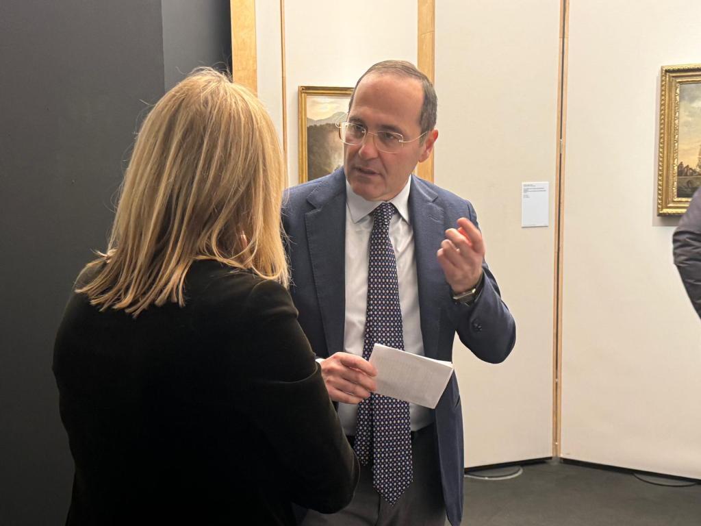 Due persone conversano in un ambiente espositivo, con opere d'arte sullo sfondo.