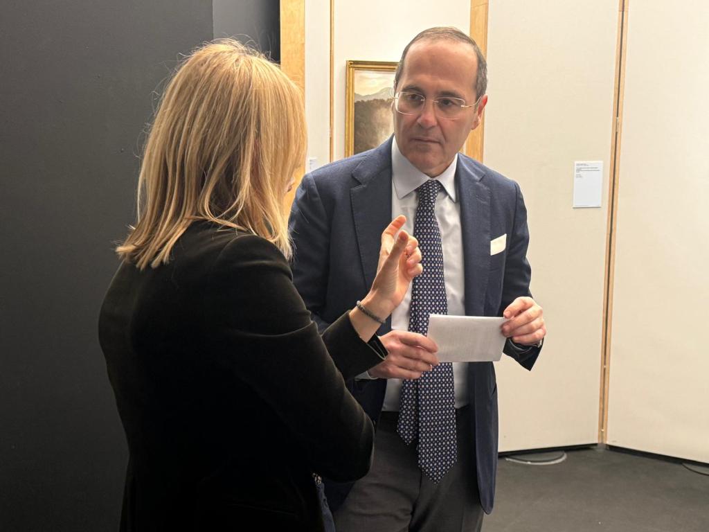 Due persone, un uomo e una donna, stanno conversando in un ambiente espositivo, con opere d'arte in vista sullo sfondo.