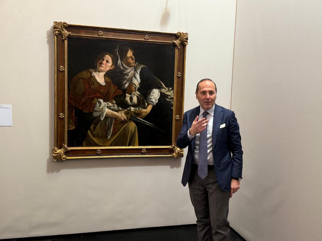 A man in business attire stands beside a large, framed painting in an art gallery. The painting features two figures in dramatic poses, with one holding a sword and the other looking distressed.