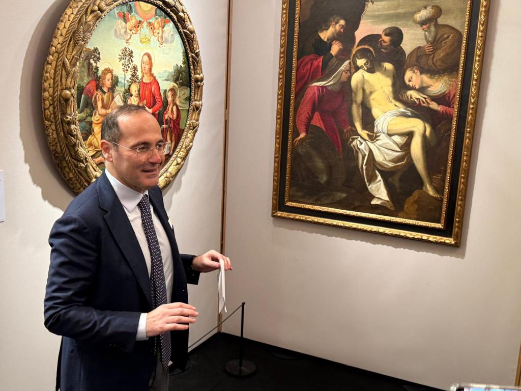 A man in a suit stands in front of two paintings in an art gallery. One painting is of the Madonna and child, while the other depicts a scene with figures around a crucified Christ.