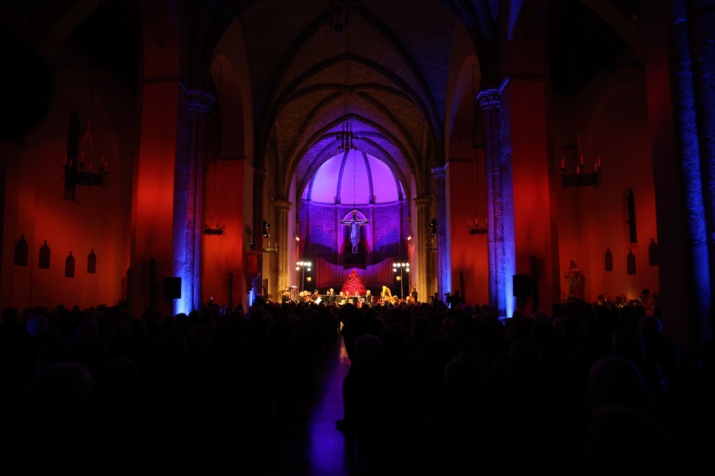 Concert taking place in a dimly lit church, featuring colorful lighting in red and blue hues illuminating the interior, with a large audience seated and a musical ensemble performing on stage.