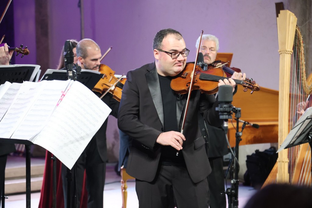 A violinist in formal attire plays on stage, surrounded by other musicians, with music sheets visible in the foreground.