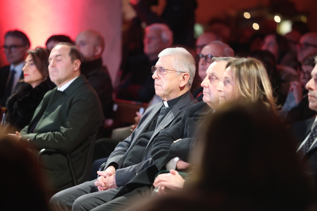 A group of people seated in an audience, some looking attentively at a speaker or event, with soft focused lighting in the background.