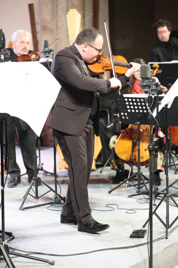 A violinist performing on stage while an orchestra plays in the background, with sheet music stands and musicians visible.