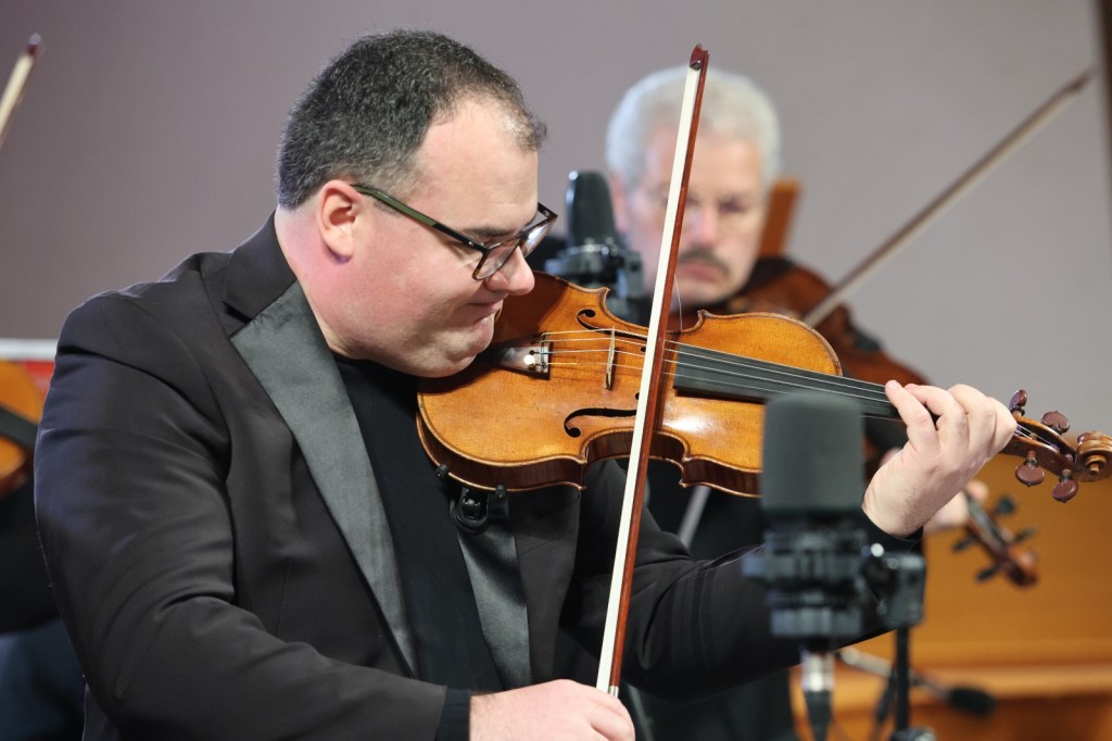 A man playing the violin with concentration, dressed in a black suit, with another musician visible in the background.