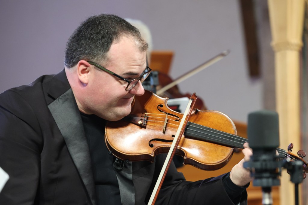 A musician playing the violin with a focused expression, wearing a black suit and glasses.